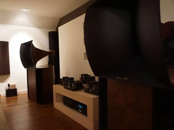 A high-end audio room featuring large Vista 3-Way Horn Speakers and a collection of tube amplifiers and preamplifiers on shelves, set in a dimly lit room with wooden floors.