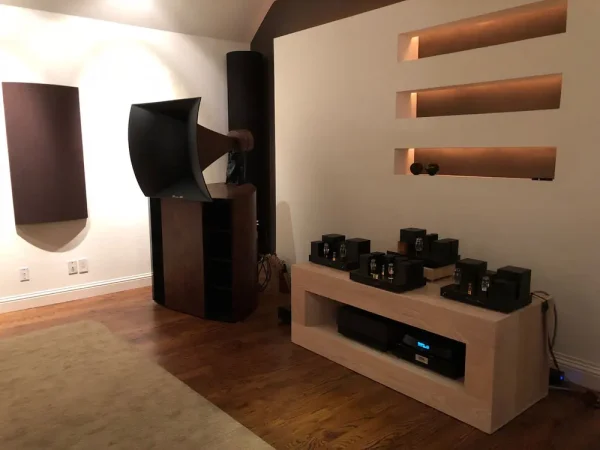 A minimalist living room featuring large Vista 3-Way Horn Speakers, wooden wall shelves, and a modern audio system with a preamplifier on a low console.