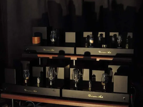 A collection of vintage tube amplifiers, including several Vista 3-Way Horn Speakers, with glowing tubes on a wooden shelf, displayed in a dimly lit room.